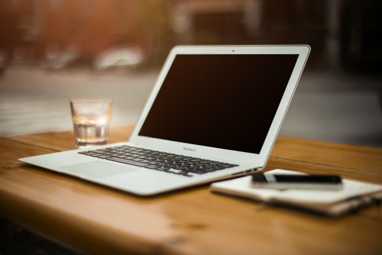 Op de foto ziet u een laptop op een houten tafel met daarnaast een notitieblok met pen een een glas water.