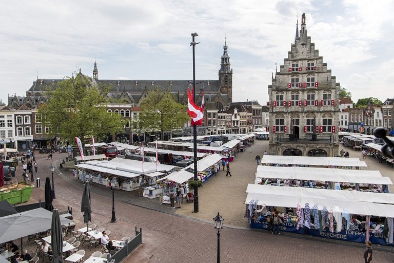 Op de foto de Markt waar de stoffenmarkt wordt gehouden. De foto is van bovenaf vanaf de Waag genomen, links verschillende terrassen met mensen en op de achtergrond het stadhuis.