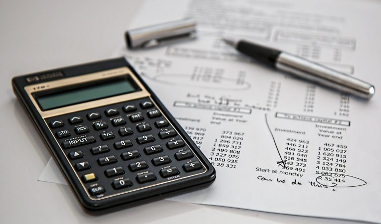 Foto van een rekenmachine op een vel papier met daarop geldbedragen en aantekeningen. Daarop ligt ook een pen.