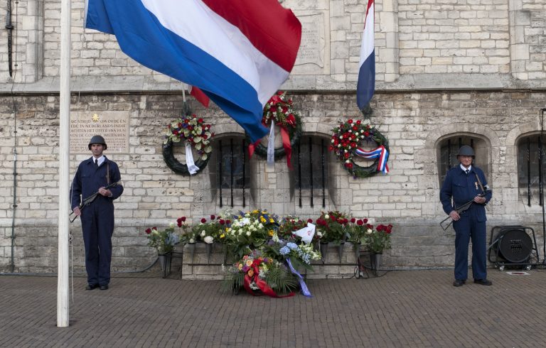 Foto van een eerdere dodenherdenking. Er hangen 3 kransen aan het oude stadhuis bij het oorlogsmonument Sint-Joris en de Draak. Er staan bloemen en 2 veteranen naast.