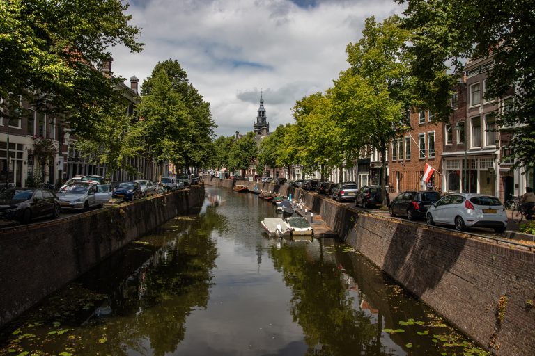 Een foto van een kanaal in de binnenstad van Gouda. Auto's en woonhuizen aan beide kanten, de Sint-Janskerk in de verte.