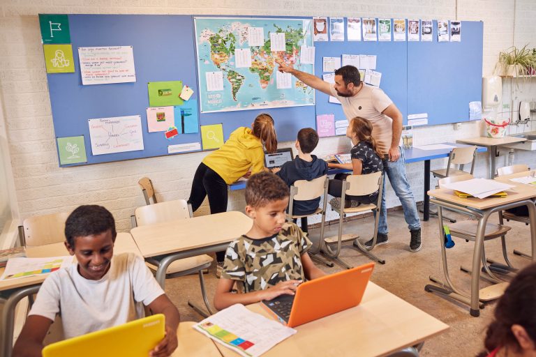 In een klaslokaal, wijst een leraar naar een blauw bord met een wereldkaart erop, drie kinderen kijken ernaar. Twee andere kinderen in de voorgrond zijn bezig op hun laptops.