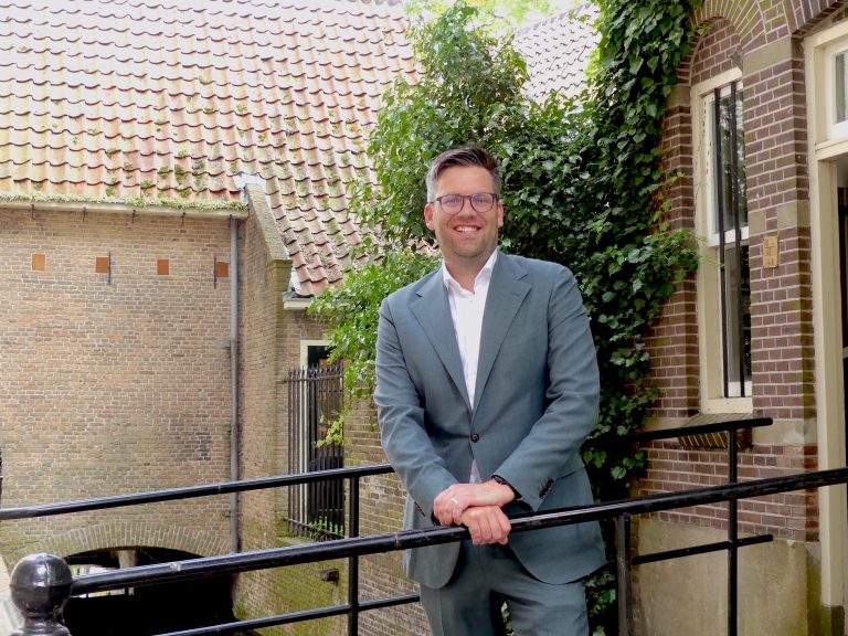 Op de foto staat wethouder Jan Kees Oppelaar op een bruggetje voor een gebouw achter de Sint Janskerk.