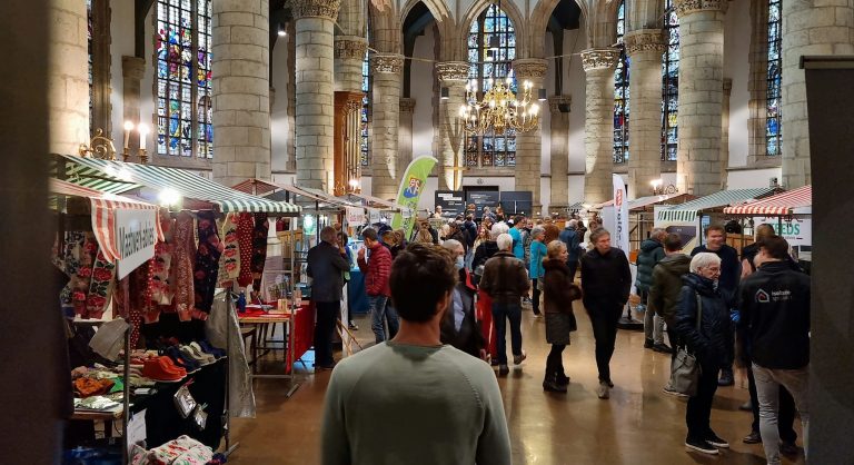 Op de afbeelding zien we de binnenkant van de Sint-Janskerk, met veel grote glas-in-lood ramen. De kerk staat vol met mensen en kraampjes. Het is een druk bezochte markt.