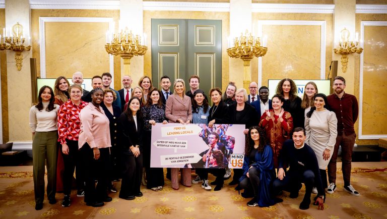 Koningin Máxima samen met vertegenwoordigers van gemeentes (wethouders, beleidsmakers en jongeren/boegbeelden). Máxima heeft een bord vast waarop staat: "Mind us leading locals, samen op weg naar een integrale aanpak van de mentale gezondheid van jongeren"