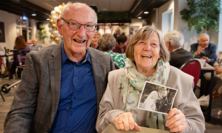 Op de foto ziet u een echtpaar dat 60 jaar getrouwd is. De vrouw heeft een trouwfoto in haar hand.