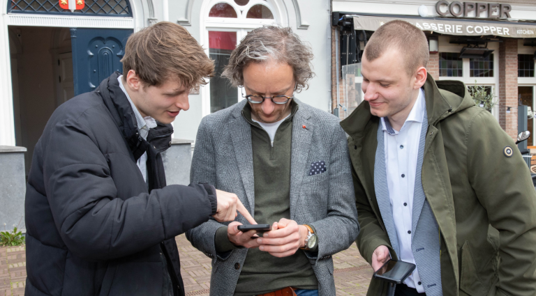 Op de foto staat Thierry van Vugt tussen de 2 ontwikkelaars van de nieuwe webapp, terwijl ze naar een telefoon kijken.