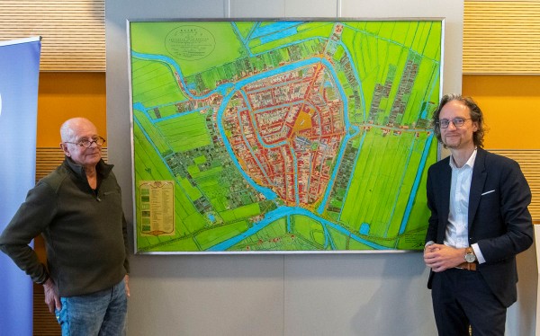 Op de foto staan links kunstenaar Sjaac van Esschoten en rechts wethouder Thierry van Vugt. Zij staan in het Huis van de Stad. In het midden hangt de stadsplattegrond van Gouda in het jaar 1828.