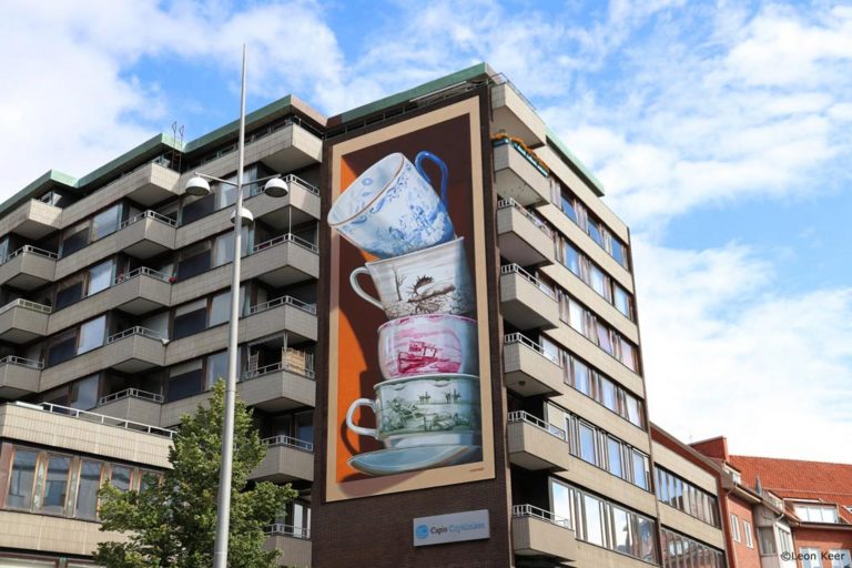 Op de foto staat een flat in de Zweedse Helsingborg met een muurschildering. De muurschildering is een optische illusie, waarbij 4 theekopjes op elkaar gestapeld staan, waarbij het lijkt alsof het bovenste kopje bijna van de stapel valt.
