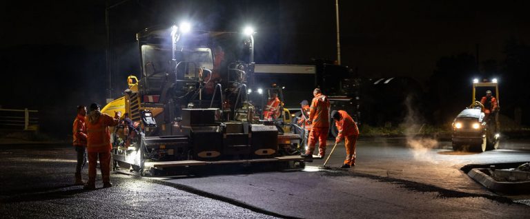 Op de foto zien we een asfalteermachine en mensen die aan het werk zijn aan de Steve Bikobrug.