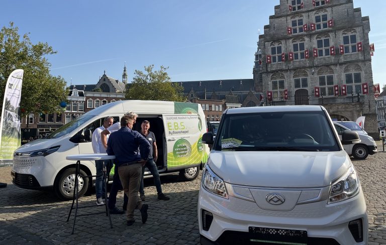 Op de foto 2 elektrische bedrijfsauto's op de Markt met op de achtergrond het stadhuis. Tussen de 2 auto's staan 3 mannen te praten aan een statafel.