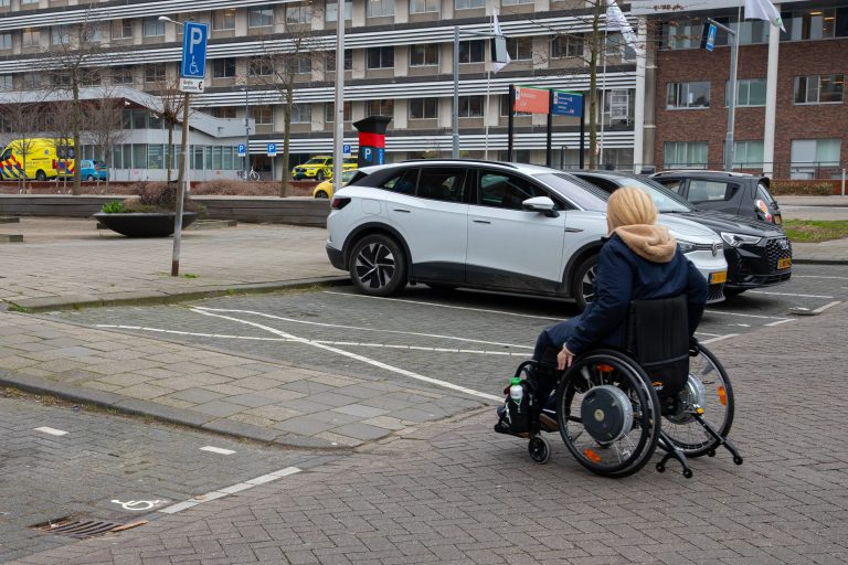 Persoon in rolstoel die een verlaagde stoep bij een parkeerplaats op rijd. De persoon in de rolstoel is warm gekleed en heeft een muts op.