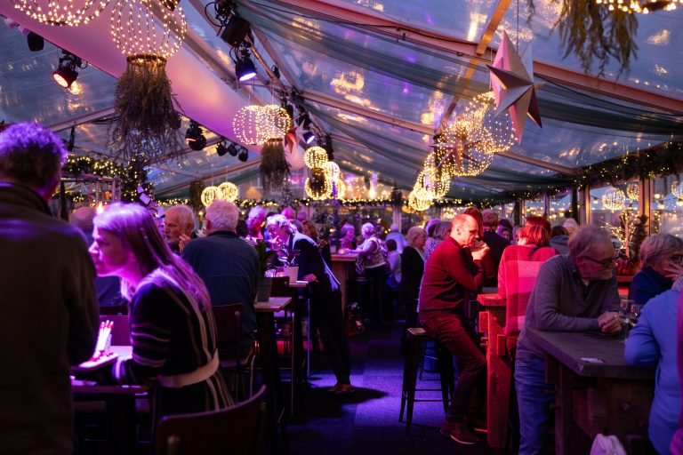 Foto van de bijeenkomst in de koek & zopie-tent op de Goudse ijsbaan. Op de foto staan allerlei bezoekers.