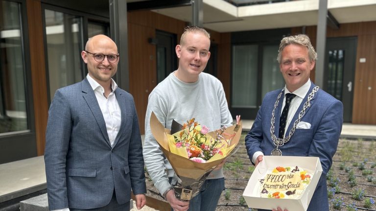 Op de foto staan drie personen afgebeeld: Bart Post (hoofd dienstverlening), inwoner Stef Ottens en burgemeester Pieter Verhoeve. Stef heeft een bos bloemen in zijn handen en de burgemeester een taart.