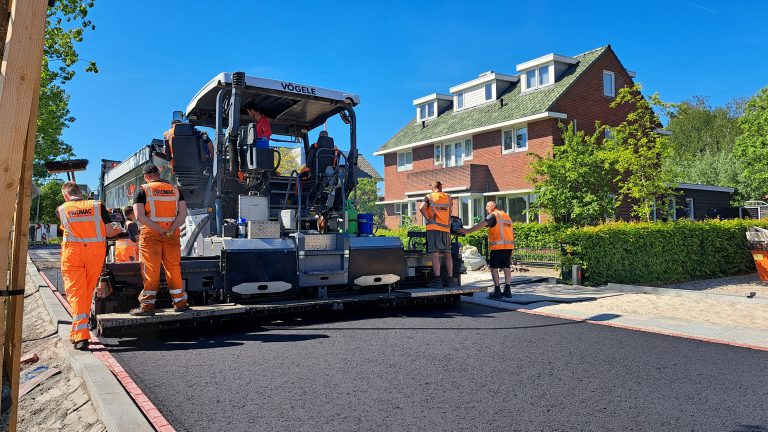 Op de foto is een grote asfalteermachine te zien die een nieuwe asfaltlaag heeft gelegd. Op en rondom de machine staan bouwvakkers met oranje pakken aan.
