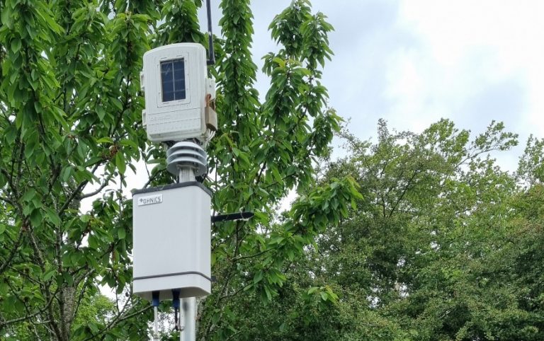 Op de afbeelding staat een fijnstofmeter. Deze hangt aan een paal in de buitenlucht.