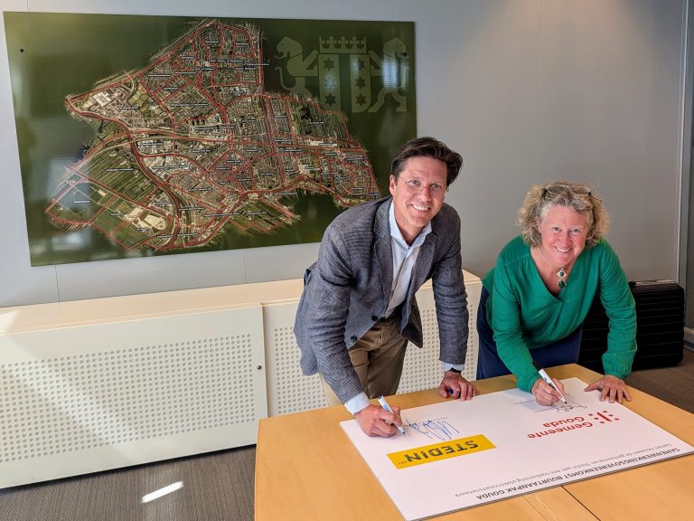 Op de foto is te zien dat Maarten Bijl, regiodirecteur van Stedin, en wethouder Judith Sargentini de samenwerkingsovereenkomst over de uitbreiding van het Goudse elektriciteitsnetwerk aan het tekenen zijn. Ze leunen voorover op tafel. Op de achtergrond hangt een luchtfoto van Gouda aan de muur.