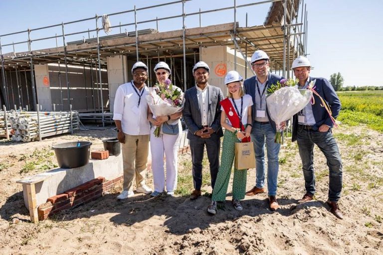 Op de foto zijn van links naar rechts te zien: twee toekomstige bewoners, Niraj Sewraj (BPD), kinderwethouder Jinthe Seppenwoolde en wethouder Jan Kees Oppelaar (gemeente Gouda) en Frans Vink (Frans Vink & Zonen). Alle mensen op de foto dragen een witte bouwhelm en een bewoner en de wethouder hebben een bos bloemen in hun hand. Op de achtergrond is een woning in aanbouw te zien.