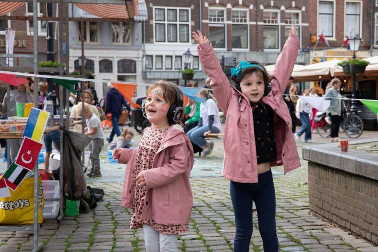 Op de foto staan twee meisjes met een roze jas aan. Een van de meisjes juicht met haar handen in de lucht
