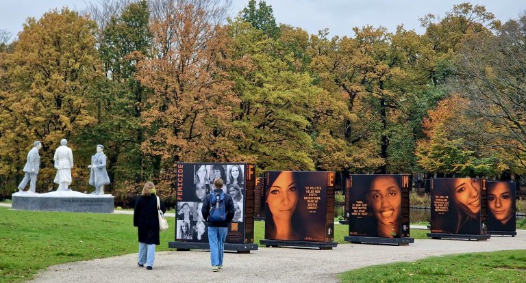 Op de foto is de buiten-expositie ‘Femicide’ te zien in een park. Er staan 5 grote borden met foto's van vrouwen.