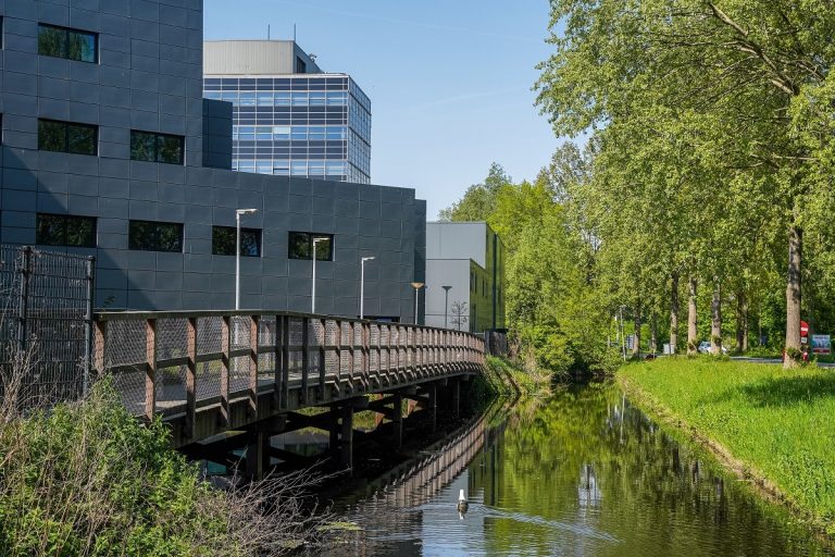 Op de foto is het gebouw van het AZC Molenwiek te zien. Naast het gebouw ligt een lange loopbrug met een sloot ernaast.