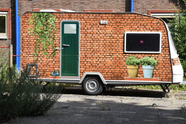 Een caravan. De kant waarop je de caravan binnen komt, is versierd met een soort van plakplastic en lijkt op de ingang van een huis. Je ziet een stenen muur, een huisdeur en planten tegen de gevel