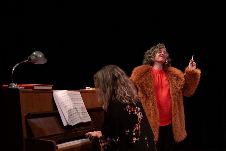 Op de foto een scène uit de voorstelling Geen vaarwel. Er speelt een vrouw op de piano. Naomi Inez staat ernaast. Ze draagt een bontjas en heeft een sigaret in haar hand.