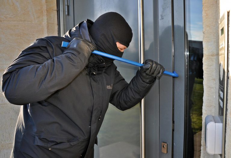 Een man met donkere kleding en een bivakmuts op heeft een koevoet in zijn hand. Hij probeert hiermee een deur open te krijgen.