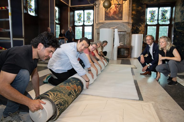 Onder toeziend oog van wethouder Thierry van Vugt en directeur Femke Haijtema van Museum Gouda wordt het koninklijk wandtapijt in de trouwzaal voorzichtig opgerold.
