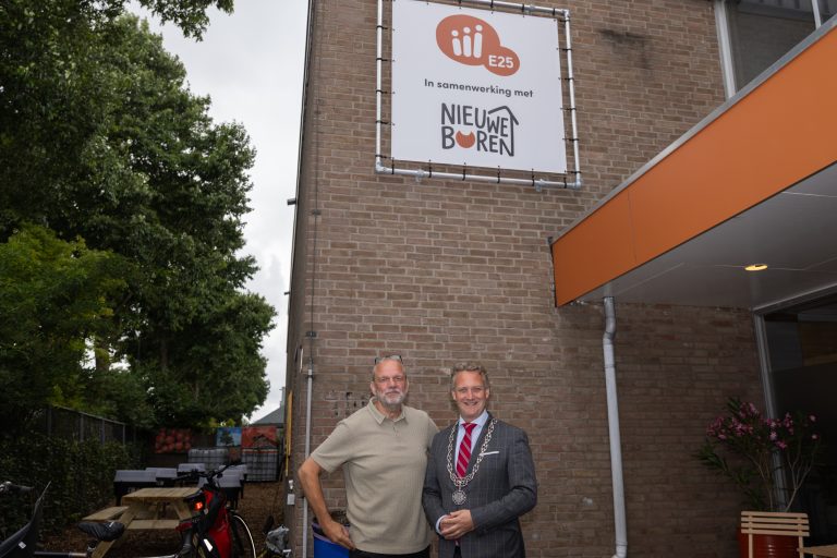 De burgemeester en een man staan voor een gebouw. Boven hen staat een bord met de tekst: E25 in samenwerking met Nieuwe Buren