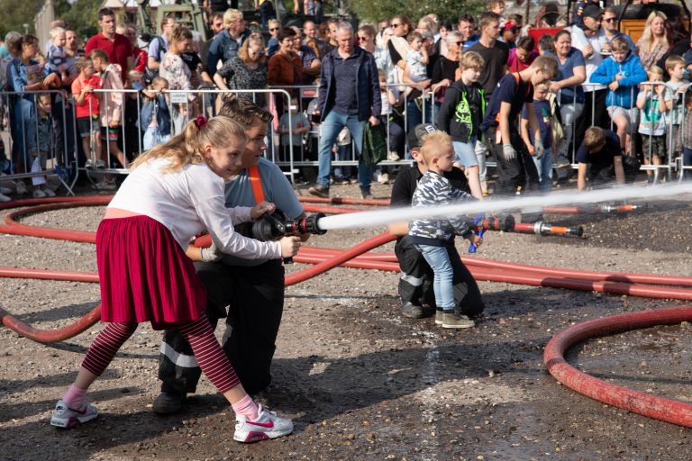 Op de achtergrond staat een groep mensen achter hekken. Op de voorgrond staan twee kinderen die ieder met een vrijwilliger van de 112 dag een water spuitende brandslang vast hebben.