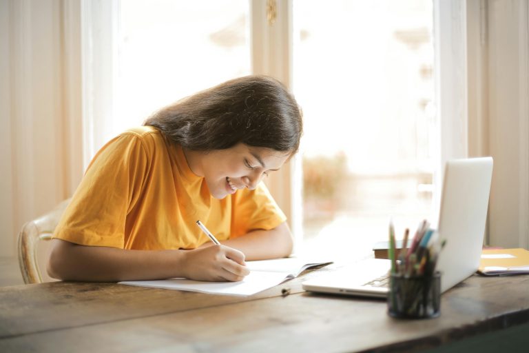 meisje zit aan tafel te schrijven met een computer voor zich.