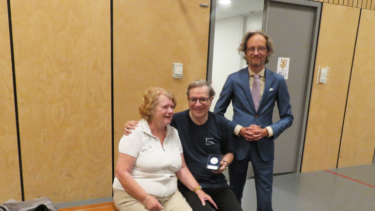 Joop Oosteen houdt de erepenning vast. Hij staat samen met zijn vrouw en Thierry van Vugt op de foto in de gymzaal.
