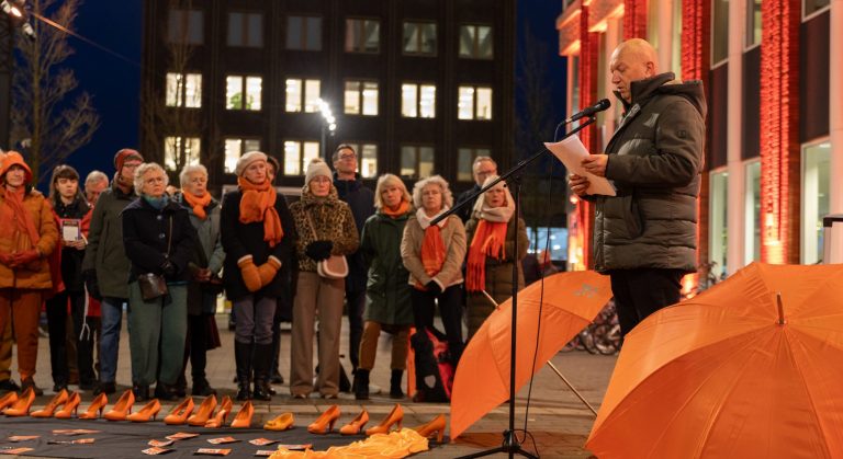 Wim Hertgens spreekt een menigte toe voor Orange the World voor het Huis van de Stad. Voor hem staan een heleboel oranje schoenen. Het huis van de stad is verlicht met oranje verlichting.