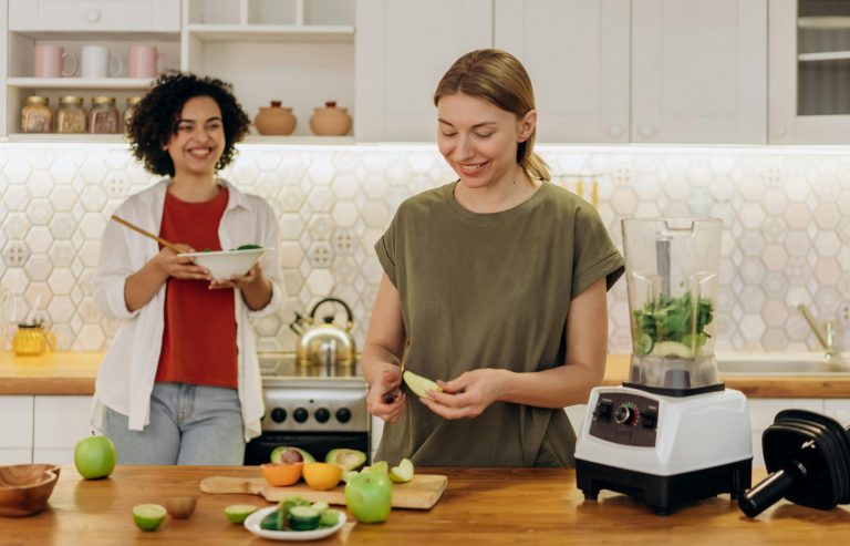 2 dames staan in de keuken. Een dame schilt een appeltje, de andere houdt een schaal eten in haar handen.
