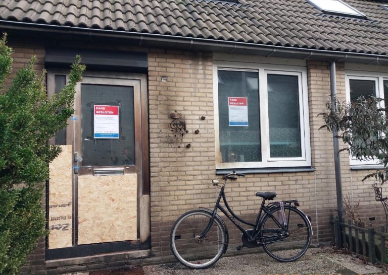 Op de foto staat een huis waarvan de deuren zijn dichtgemaakt met hout. Op de deur en het raam staat een aanplakbiljet met de woorden 'Pand gesloten'. Er staat een fiets voor het huis.
