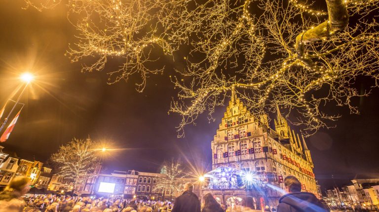 De binnenstad tijdens kaarsjesavond. Het is donker, mensen staan rondom het stadhuis en er brandt veel licht.