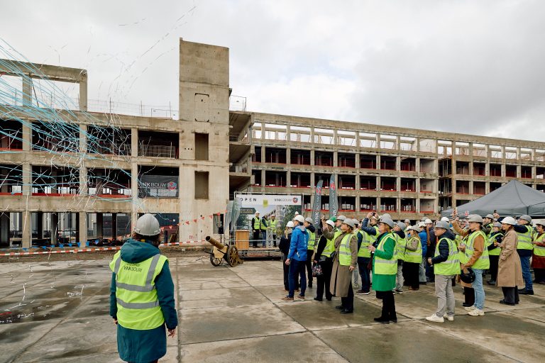Op het bouwterrein van de Kazerne wordt er confetti uit een kanon geschoten. Er staan een groep mensen met gele hesjes en witte helmen bij om het moment te vieren.