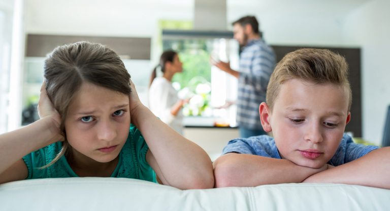 Afbeelding van 2 jonge kinderen die op de bank zitten. Op de achtergrond hebben hun ouders ruzie.