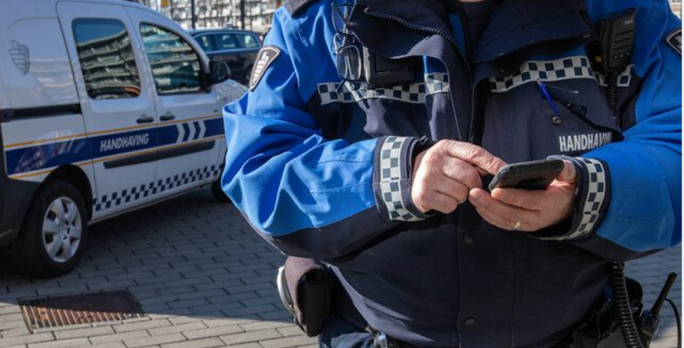 Close-up van de handen van een man in een blauw uniform: hij registreert iets op een zwart apparaat. Op de achtergrond een auto met 'Handhaving' op de zijkant.