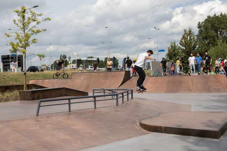 Een skateboarder springt van een rail in het nieuwe skatepark. Er staan een aantal mensen te kijekn en op de achtergrond staat een bmx-er.
