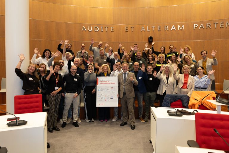 Gouda is gestart met een Lokale Zorg- en Veiligheidskamer. Een foto in de raadzaal van het Huis van de Stad, waar medewerkers van de gemeente en verschillende zorg-, veiligheids- en maatschappelijke organisaties op 13 oktober hiervoor een convenant tekenden. Iedereen zwaait naar de camera en burgemeester Pieter Verhoeve houdt het convenant vast.